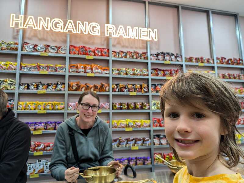 Sonia and Jake sitting in front of a wall of 2 minute noodles